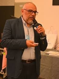 Man with glasses and beard in dark blazer holding microphone while speaking at an indoor event